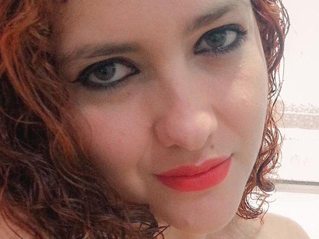 Das Gesicht von GinebraNova mit lockigem, rotem Haar, blauen Augen, schwarzem Eyeliner und rotem Lippenstift, zeigt ein leichtes Lächeln und eine kleine Nasenpiercing. Ihre Haut ist hell und leicht glänzend..