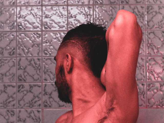 AngelsScott has a shaved side, raised arm, and visible armpit hair in a red-lit photo, showing a muscular, bearded neck and profile against a textured wall.