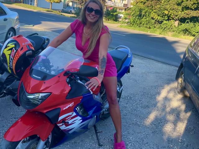 LaMadrina with long blonde hair, wearing a pink dress and matching heels, leaning on a red and blue Honda sport bike, smiling, with a helmet on the left.