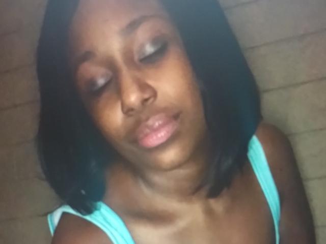 SexySexyMymy with dark skin, black shoulder-length hair, and pinkish lips, wearing a turquoise top, eyes closed, and a slight smile, in a slightly blurry photo.