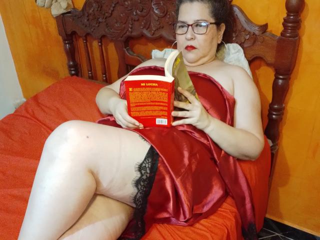 Loretta Sanchez, eine etwas mollige Frau mit Brille und rotem Lippenstift, liegt auf einem Bett aus rotem Satin, schwarzer Spitze und einem Buch, wobei ihre helle Haut und ihre Oberschenkel sichtbar sind..