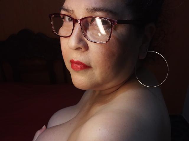 Loretta Sanchez, avec du rouge à lèvres, des lunettes et des boucles d'oreilles pendantes, a ses seins exposés, une peau claire et des cheveux foncés, dans une pièce faiblement éclairée, et regarde légèrement sur le côté..