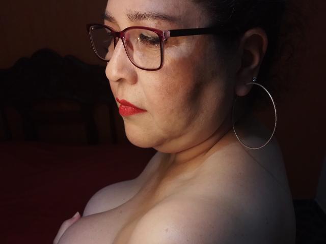 LorettaSanchez, with red lipstick and glasses, has her tits exposed, wearing large hoop earrings, and a slightly sad expression, in a dimly lit, brown-toned room.