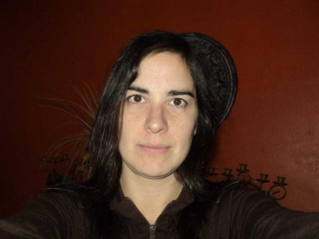 EstrellaHott, con lunghi capelli neri, occhi marroni e un'espressione neutra, indossa una giacca marrone scuro con zip, davanti a un muro rosso, con un cappello nero leggermente inclinato..