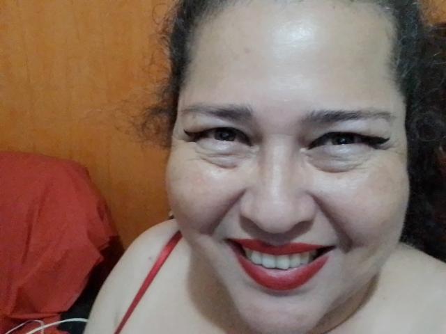 LorettaSanchez has curly dark hair, red lipstick, and black eyeliner, smiling with slightly wrinkled eyes, wearing a red strap, looking happy.