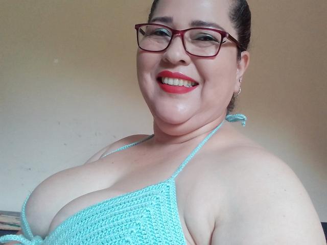 Loretta Sanchez, com corpo robusto e seios grandes, vestindo um top de crochê azul, sorrindo, usando óculos de armação vermelha, com cabelo escuro preso em um coque, pele clara e batom vermelho..