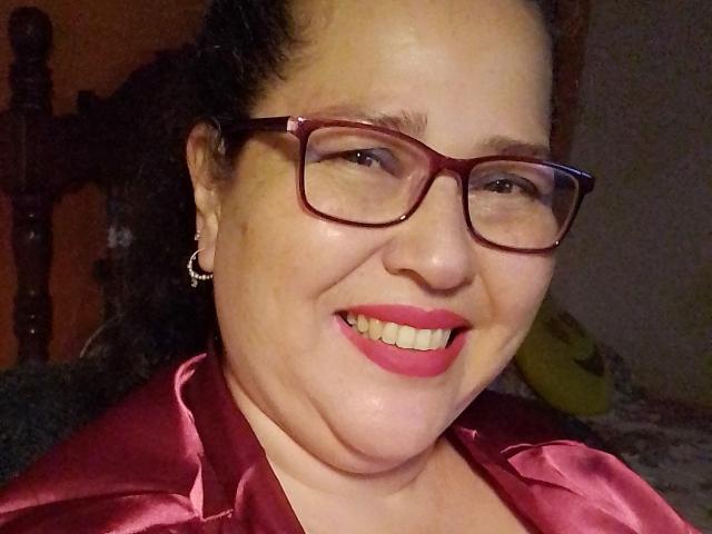 Loretta Sanchez, met rode bril, roze lippenstift en een glanzend rood satijnshirt, glimlacht naar de camera, toont haar mollige gezicht en draagt kleine oorringen..