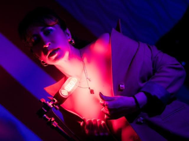 ViktoriaBrown with short brown hair, wearing a pinkish-brown blazer, red earrings, and a necklace, under purple and pink lights, holding a microphone, her tits slightly visible.