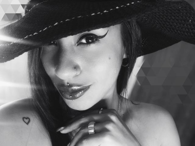 LunaCandy, con un sombrero negro, lápiz labial oscuro y un piercing en la nariz, mostrando un tatuaje de corazón en el hombro, llevando un anillo en el dedo y con un delineado de ojos llamativo..