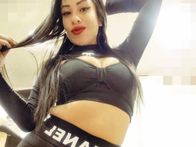 LunaCandy, con cabello largo y negro, lápiz labial rojo y un top deportivo negro que deja ver sus senos, levantando su brazo derecho, vestida con pantalones negros "JAWA", posando con confianza..