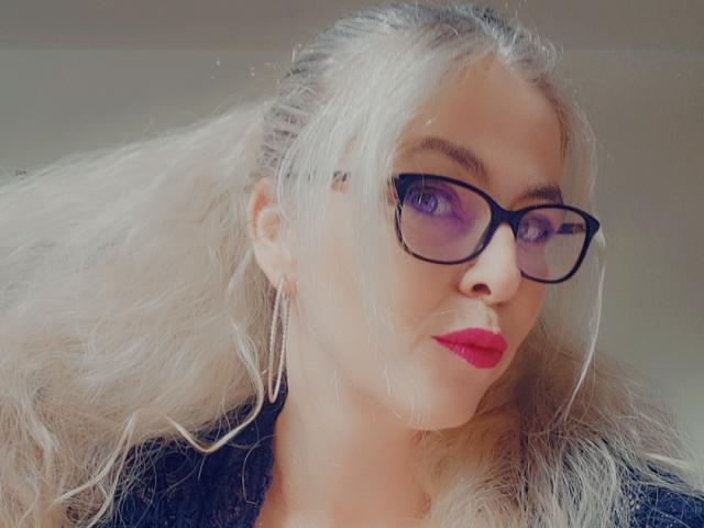 Melusia, com cabelo loiro, batom vermelho, óculos pretos e brincos de argola, exibindo seus seios em uma selfie levemente desfocada. Seu traseiro não está visível..