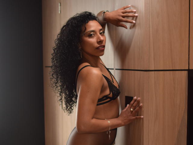 JanEvans, com cabelo preto encaracolado, vestindo um sutiã e calcinha de renda pretos, apoiada em uma parede de madeira, olhando por cima do ombro, com as mãos na parede, pele morena, seios pequenos, sexy, pulseira no pulso..