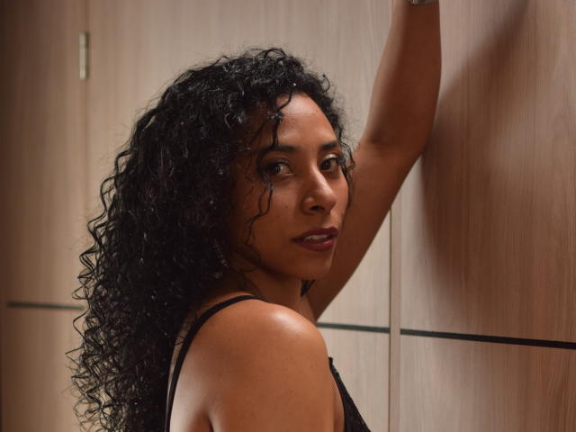 Jan Evans, met krullend zwart haar, bruine huid en donkere ogen, draagt een zwart tanktop, met een arm omhoog, kijkt over haar schouder, met een subtiele glimlach, heeft een gemiddelde borstmaat, met een houten achtergrond..