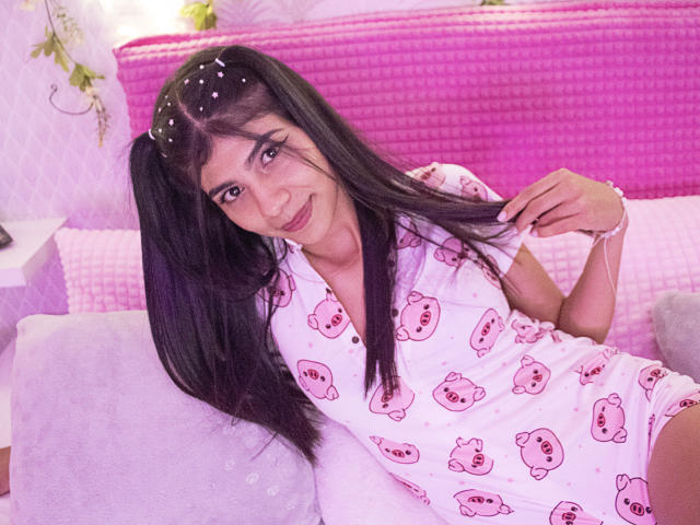 SamyNicols, com cabelos longos e pretos, vestindo uma camisa de pijama rosa com estampa de ursos, sorrindo, segurando um tufo de cabelo, usando uma tiara rosa, em uma cama rosa, com uma parede branca, fofa, sexy, relaxada..