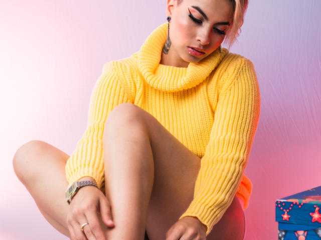 CloeBransoon in a yellow rib-knit sweater, crouched with one knee up, showing her bare ass. She has pink eyeshadow, a tattoo, and a bracelet, looking down.