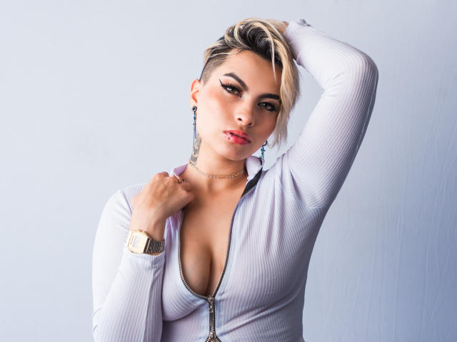 Cloe Bransoon, avec ses cheveux blonds courts, porte un haut blanc côtelé à fermeture éclair qui expose sa poitrine. Son bras est levé, elle porte une montre en or, un collier en chaîne et du rouge à lèvres. Elle a une allure séduisante..