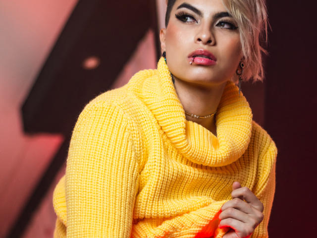 Cloe Bransoon met kort blond haar, een gele gebreide trui, roze lippenstift, een neusring, die naar de zijkant kijkt, een oranje papiertje vasthoudend, donkere wenkbrauwen, subtiele make-up, sexy, zelfverzekerd..