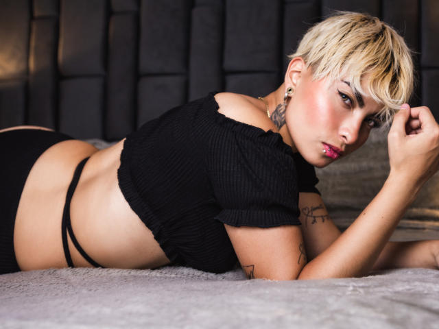 Cloe Bransoon, con i capelli biondi corti, sdraiata su un letto, indossa una maglia nera a coste e uno slip nero, mostrando il sedere e i tatuaggi, e guarda verso la fotocamera..