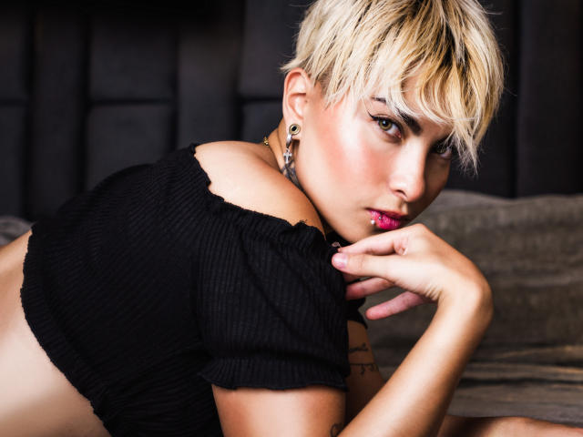 Cloe Bransoon, con cabello rubio corto, una blusa negra con un hombro descubierto y lápiz labial rosa, está acostada en una cama, mirando a la cámara con la mano cerca de su boca, mostrando un tatuaje..