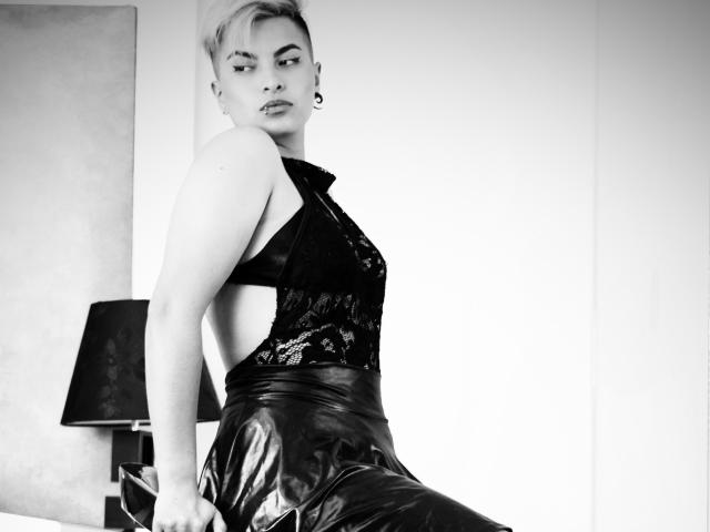 CloeBransoon with short blonde hair, lace-up black top, and shiny black leather skirt, sitting on bed, looking over shoulder, black lamp in background, black and white photo.