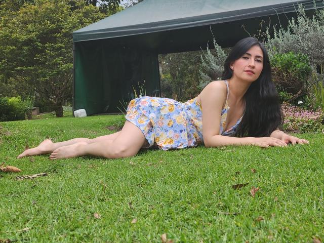 JainaRogers, lying on grass, legs stretched out, wearing a floral dress, barefoot, black hair, looking at camera, green shed in background, cute, small tits, pretty, relaxed.