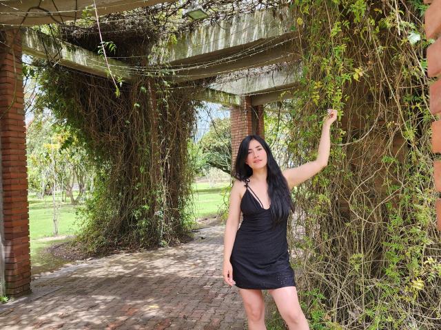 JainaRogers with long black hair, wearing a black dress, standing under a brick and vine-covered structure, raising her right arm, showing a slight smile, with green grass behind.