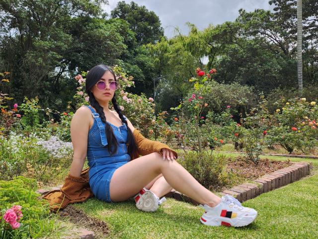 JainaRogers in een blauwe spijkerjurk, zittend op het gras met gekruiste benen, met roze zonnebril, witte sneakers en een bruine trui, met lange zwarte vlechtjes, in een rozen tuin..