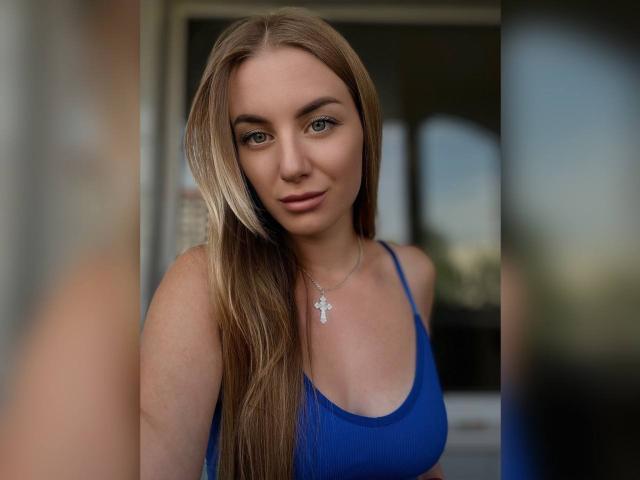 BlondyOneSex mit langen blonden Haaren, einem blauen Sport-BH und einer silbernen Kreuzkette, steht drinnen und schaut mit einem leichten Lächeln in die Kamera. Ihre Brüste sind sichtbar..