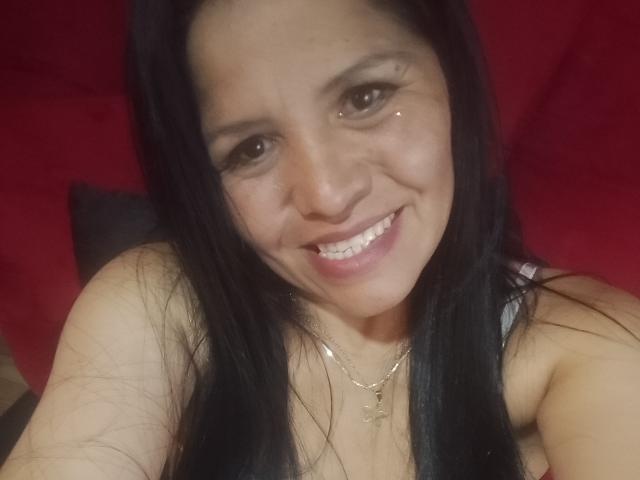 Melanie69 has long black hair, a smile showing white teeth, and a gold cross necklace, with a small sparkle on her right eyelid. She's wearing a sleeveless top.