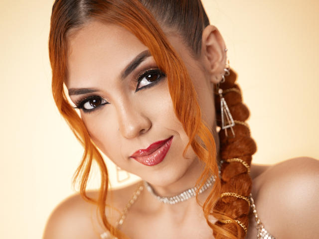 Maisy Warner, met oranje gevlochten haar, donkere eyeliner, rode lipstick, glanzende oorbellen en een sierlijke ketting, glimlacht naar de camera en laat haar sexy, aantrekkelijke gezicht zien..