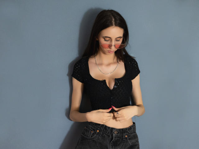 KatArt, avec de longs cheveux bruns, portant un haut noir à manches courtes et à boutons, qui révèle ses petites seins, et un jean foncé, utilisant un téléphone rose devant un mur bleu..