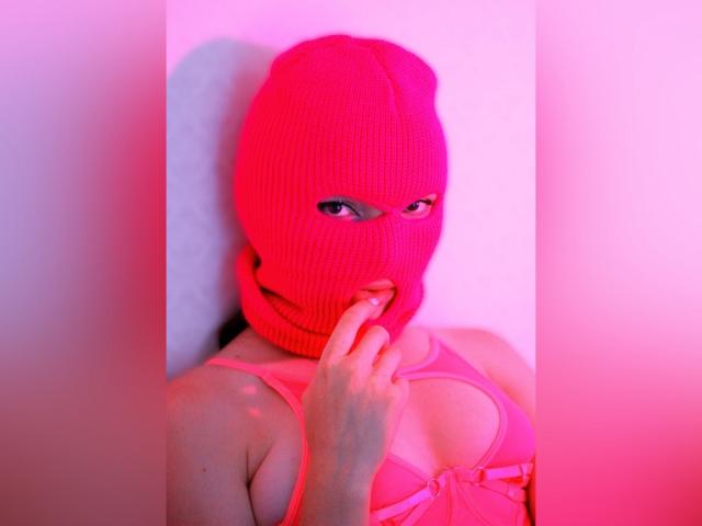 WebLux trägt eine rote, gerippte Maske, beißt sich in den Finger, hat rosafarben beleuchtete Haut und einen rosa BH, der ihre Brüste zeigt. Ihre Augen schauen durch die Maske..