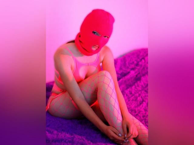 WebLux, avec un masque rose, des bas résille et un soutien-gorge rose, assise sur un lit violet, les genoux repliés, les bras autour des jambes, montrant de petites seins et un string rose, sous un éclairage rose et violet..