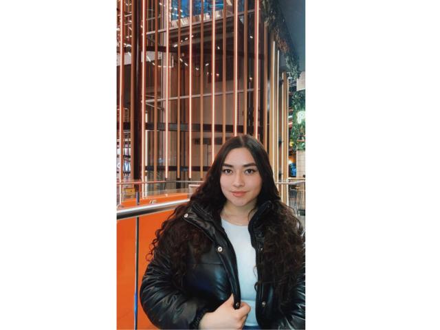 AntonnellaTyler with curly black hair, wearing a black leather jacket over a white shirt, standing in front of an orange and metal railing, looking at the camera.