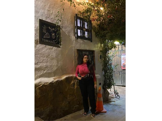 Antonnella Tyler está de pie frente a una pared áspera de color beige, vestida con una camiseta rosa de manga larga y pantalones negros, con las manos en los bolsillos, y con las nalgas ligeramente prominentes..