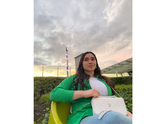 Antonnella Tyler, mit langen, dunklen Haaren, einem grünen Pullover, einem weißen Oberteil und blauen Jeans, sitzt auf einem gelben Stuhl und hält eine weiße Handtasche mit einem kleinen Logo, vor einem bewölkten Himmel..