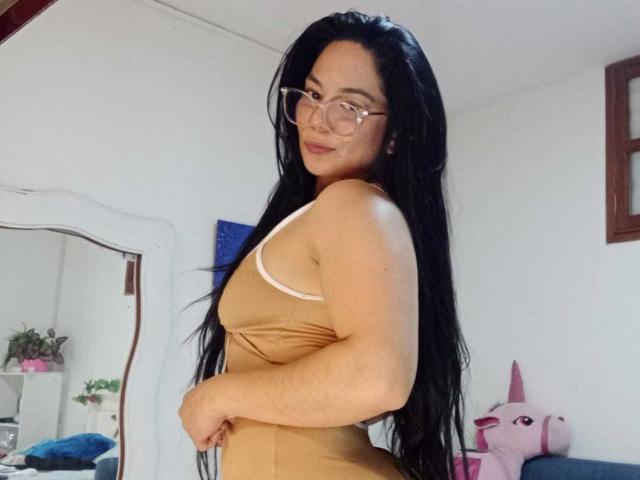 Bianca Scott mit langem, schwarzem Haar, die eine Brille trägt und ein eng anliegendes, beiges Kleid trägt, das ihr Gesäß und ihre kleinen Brüste zeigt, steht in einem Raum mit einem Spiegel und einem rosa Einhorn..