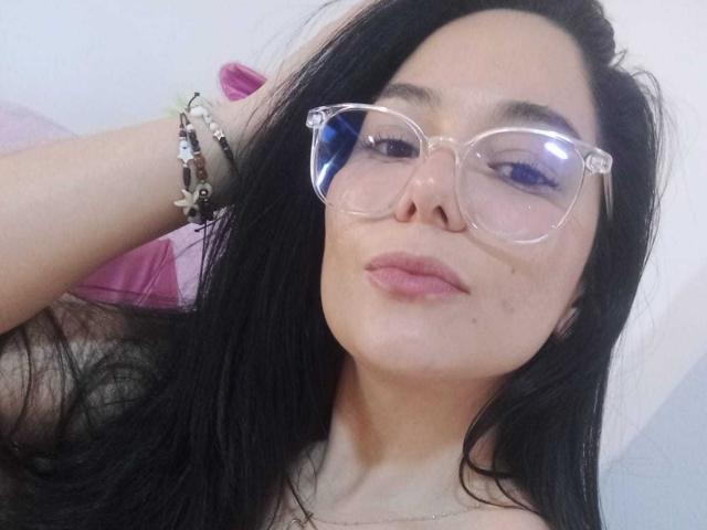 Bianca Scott, con cabello negro, gafas claras y labios rosados, lleva una pulsera y un collar, y tiene un rostro ligeramente sonrojado con una pequeña imperfección en la mejilla derecha..