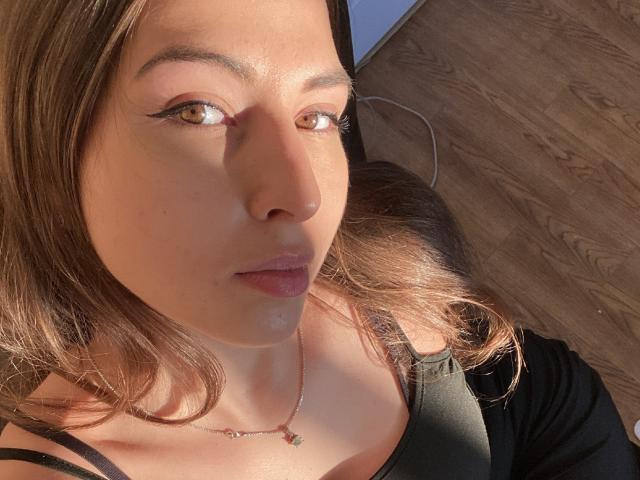DominikaRos tiene la piel de color marrón claro, ojos marrones con delineador negro y lápiz labial rosa. Lleva una blusa negra, un collar y tiene el cabello marrón claro y ondulado..