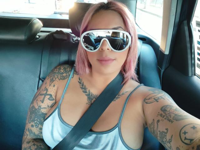 Chili, a heavily tattooed milf, wears white sunglasses, a silver tank top, and a seatbelt, taking a selfie in a car.