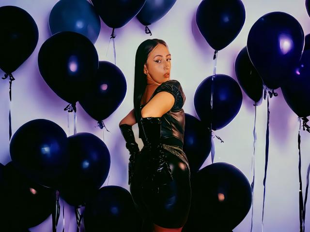 ClaireMortin, gekleed in een glanzend zwart latex outfit, zit op een motorfiets in zwart en wit, met gekruiste benen, haar hand op het stuur, en ziet er verleidelijk uit, met rode lippenstift en donker haar..