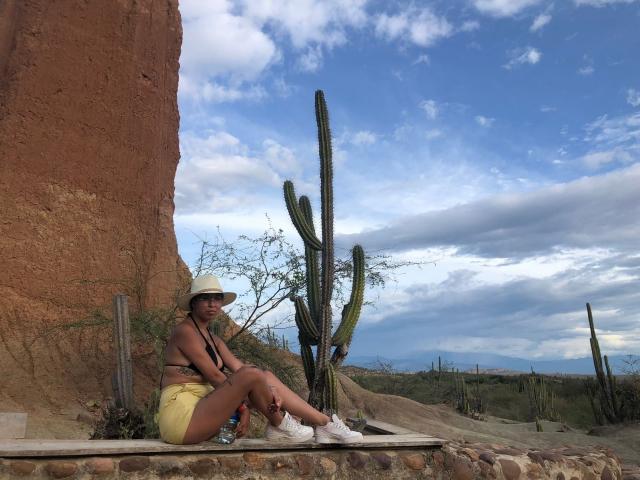 Samantha Morgan, con piel morena, busto medio, pantalones cortos amarillos, zapatillas blancas y un sombrero, sentada en un borde de piedra, al lado de cactus altos, frente a un acantilado rojo..