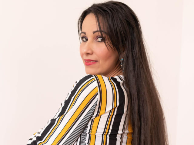LauraFoxh, com cabelo preto longo, camisa listrada amarela, branca e preta, sorrindo para a câmera, olhando por cima do ombro, usando pequenos brincos azuis, batom rosa, fundo branco..