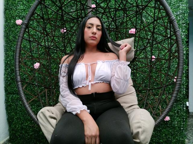 AmmyLane with black hair, white off-shoulder top showing her tits, black pants, sitting in a hanging chair, holding her hair, green wall with pink flowers in background.