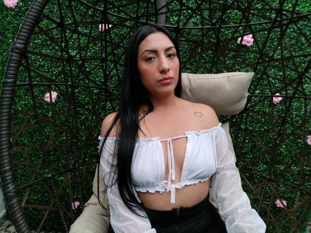 AmmyLane with long black hair, white off-shoulder top showing her tits, small heart tattoo on left shoulder, sitting in a hanging chair, green background, pink flowers.