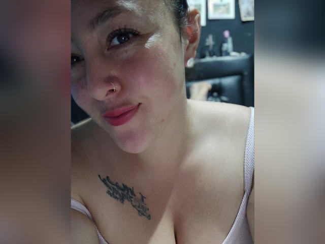 Une femme avec une peau claire, des cheveux noirs attachés en chignon, un anneau au nez, du rouge à lèvres rose et un tatouage au-dessus de ses seins. Elle porte un haut rose qui dévoile légèrement sa poitrine..