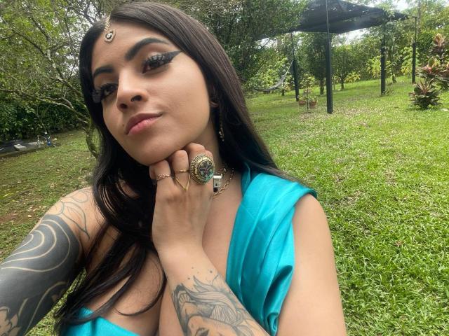 CoriPaige with dark hair, blue dress, tattoos on arm and hand, ring on finger, black eyeliner, green necklace, grassy outdoor setting, slight smile, hand on chin, selfie.