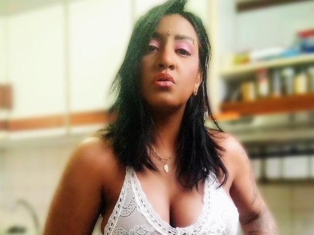Nahirr has dark hair, pink eyeshadow, and is wearing a white lace bralette that shows her tits. She's looking at the camera with a pouty expression.