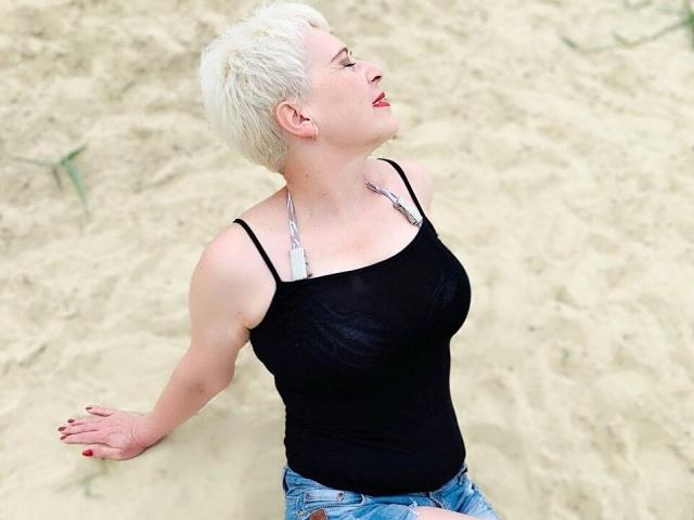 NikaClare, com cabelo loiro curto, camiseta preta e calça jeans azul, sentada no chão arenoso, com os olhos fechados, a cabeça inclinada para trás, batom vermelho, colar e esmalte vermelho nas unhas..