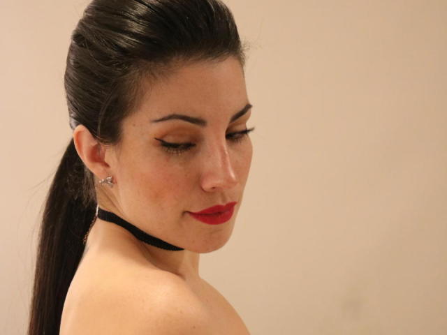 Liliysweet has dark hair in a ponytail, red lipstick, and a black choker, with closed eyes and subtle makeup, showing her bare shoulders and small earrings.